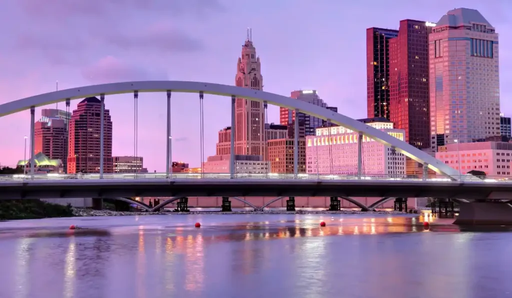 City skyline at dusk featuring a modern arched bridge over a calm river. Skyscrapers and buildings are illuminated with a pink and purple hue from the sunset, reflecting on the water.
