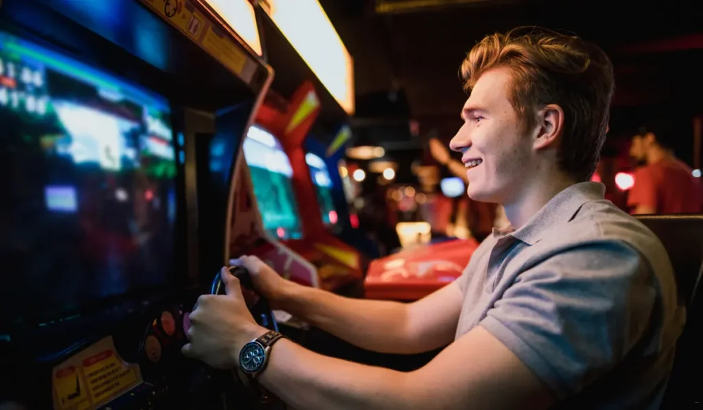 A young man with short light brown hair is smiling while playing an arcade racing game. He is holding a steering wheel, and the arcade screens are brightly lit. The background is dimly lit with some people and more arcade machines visible.
