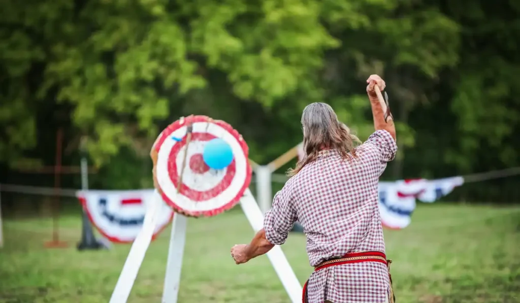 A person in a checked shirt and long hair is throwing an axe at a target painted with red and white circles. A blue balloon is attached to the target. The setting is outdoors, with green trees in the background.
