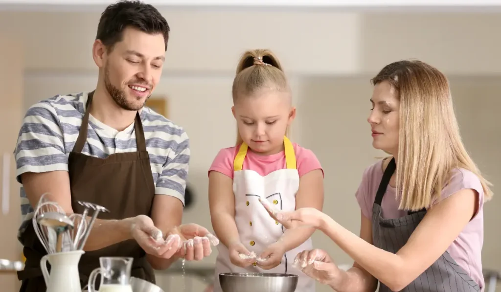 A family of three, wearing aprons, stand around a kitchen counter. The father and daughter are mixing ingredients in a bowl, while the mother looks on, guiding them. They are smiling and engaged in the cooking activity together.
