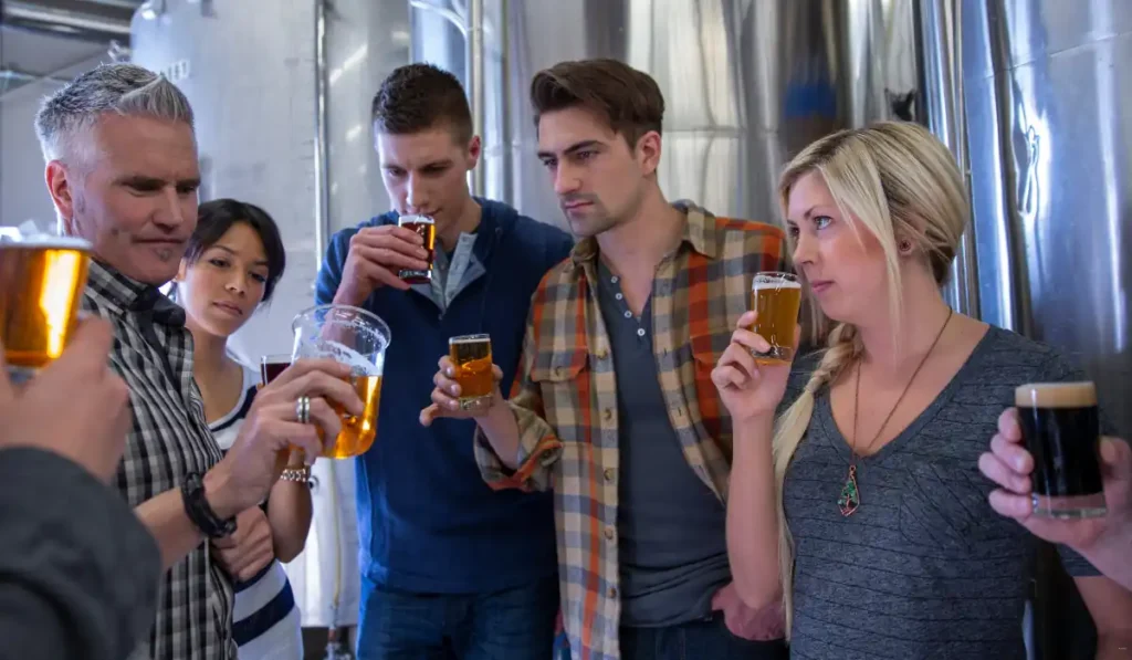 A group of people stand together in a brewery, each holding a glass of beer. They appear to be sampling and examining the beverages, with large metal tanks in the background. The atmosphere is focused and curious.
