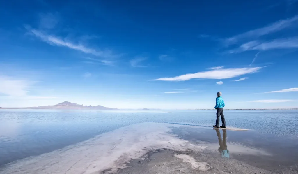 A person in a blue jacket and black pants stands on a narrow strip of land, gazing across a vast, reflective salt flat under a bright blue sky with scattered clouds. Distant mountains are visible on the horizon.
