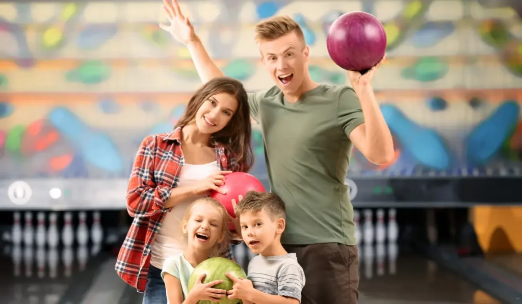 A smiling family of four at a bowling alley, each holding a bowling ball. The father and mother are standing behind their two young children. The background shows a row of bowling pins and a colorful mural.
