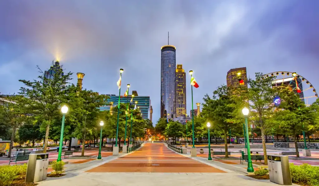 A vibrant cityscape featuring a wide, lit pathway lined with trees and lampposts leading towards tall skyscrapers. The Ferris wheel on the right adds a touch of whimsy to the urban setting under a cloudy sky.
