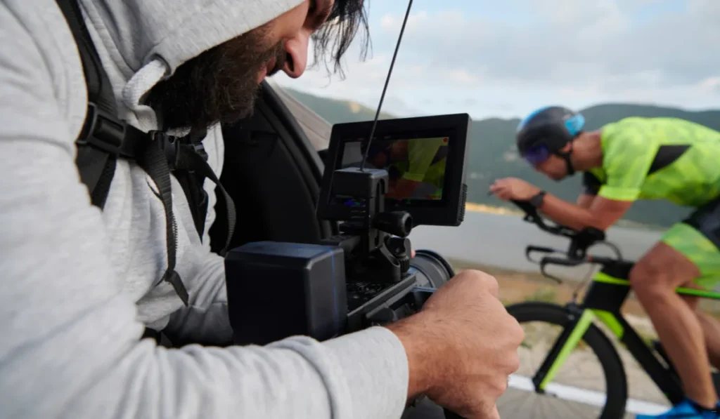 A person in a hooded sweatshirt films a cyclist in a neon green outfit riding a bike. The cyclist is racing on a road beside a body of water, with mountains in the background. The camera's screen displays the cyclist's image.
