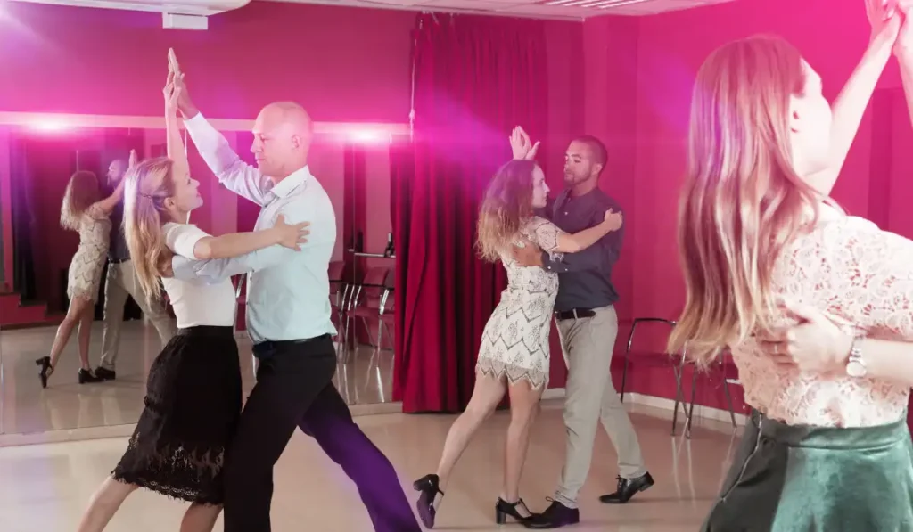 Four couples are ballroom dancing in a studio with red walls and mirrors. They are gracefully moving in pairs, with a pink light illuminating the room, and chairs visible in the background.
