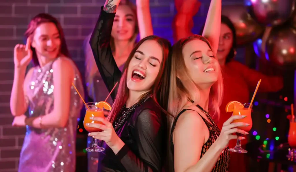 Two women with long hair are smiling and dancing back-to-back, holding orange drinks with straws. They're surrounded by others at a lively party, with colorful lights and balloons in the background.
