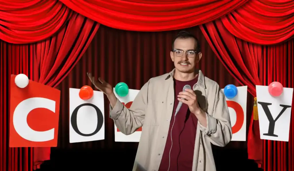 A person with a mustache and glasses is on stage holding a microphone, performing stand-up comedy. Red curtains and the word "Comedy" in colorful letters are in the background.
