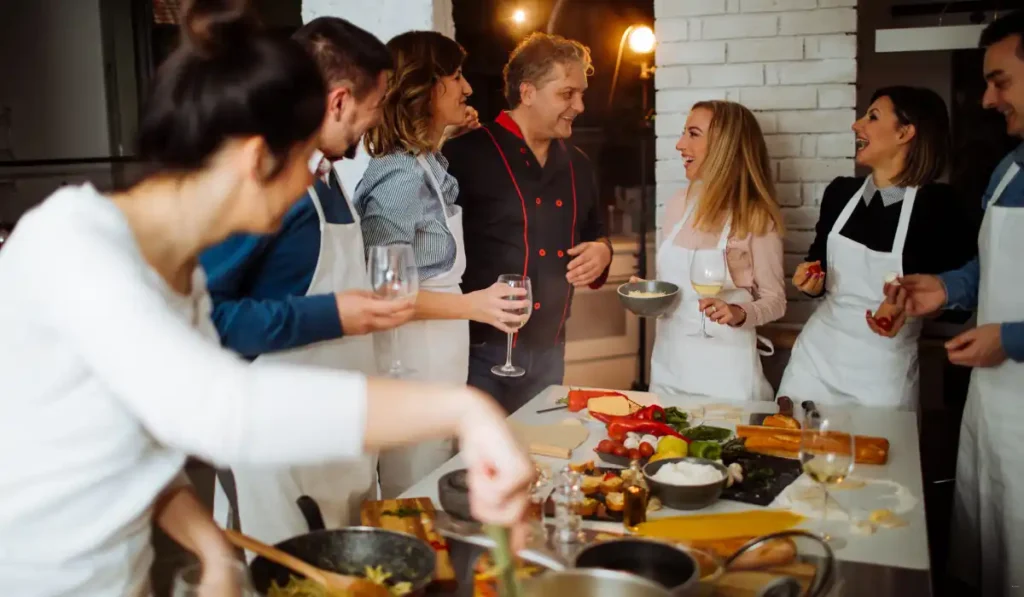 A group of people in a kitchen setting, wearing aprons, are gathered around a table filled with various ingredients and dishes. They are smiling and conversing, with one person holding a glass of wine. Cooking utensils and food items are visible on the table.
