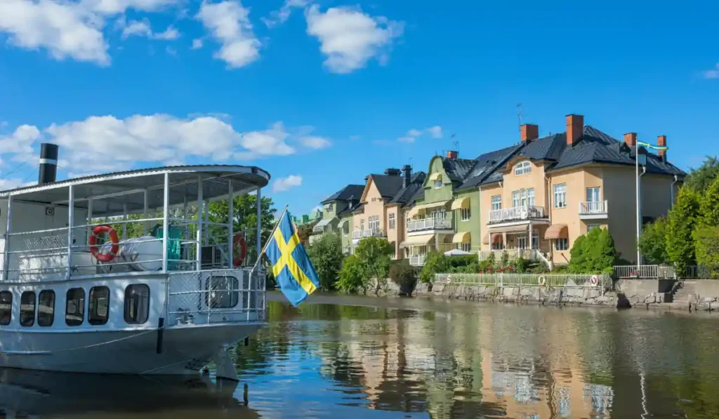 A scenic view of a river with a boat displaying a Swedish flag. In the background, colorful houses line the riverbank under a clear blue sky with scattered clouds.
