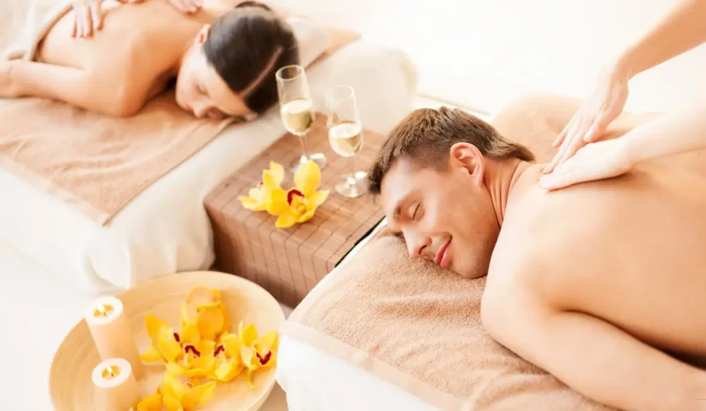 A man and a woman enjoy a relaxing massage on spa tables, each covered with a towel. Yellow orchids and two glasses of white wine are nearby, adding to the tranquil atmosphere.
