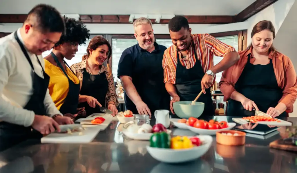 A diverse group of six people in aprons are in a kitchen, smiling as they prepare food together. Some chop vegetables on cutting boards while others mix ingredients in a bowl. Various fresh vegetables are visible on the counter.
