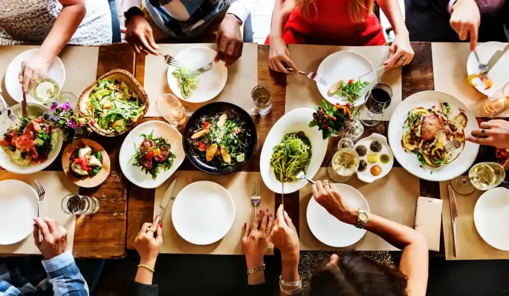 A group of people is seated around a wooden table filled with various dishes, including salads, pasta, and roasted meat. Hands are reaching for food, and there are drinks on the table, suggesting a communal meal or gathering.
