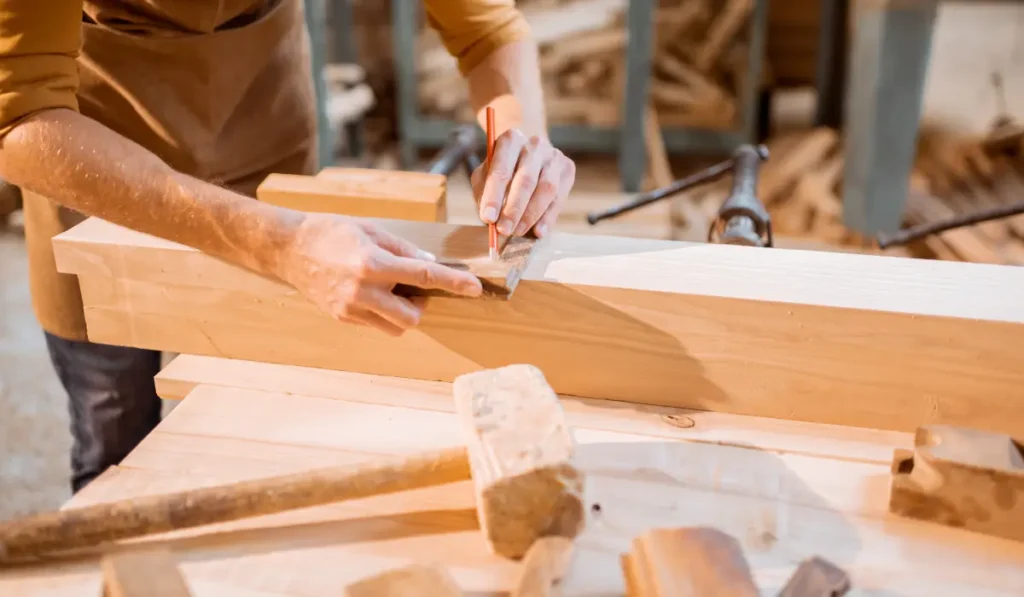 A person wearing an apron uses a square ruler and pencil to mark measurements on a wooden plank in a workshop. Various carpentry tools, such as a hammer and wood pieces, are on the workbench.
