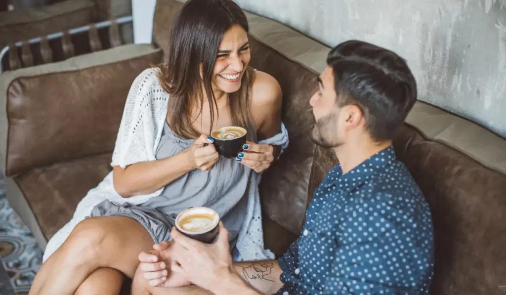 A woman and a man sit on a brown couch, smiling at each other while holding cups of cappuccino. The woman wears a gray dress and a white shawl, and the man wears a blue polka dot shirt. They appear to be enjoying a casual conversation.
