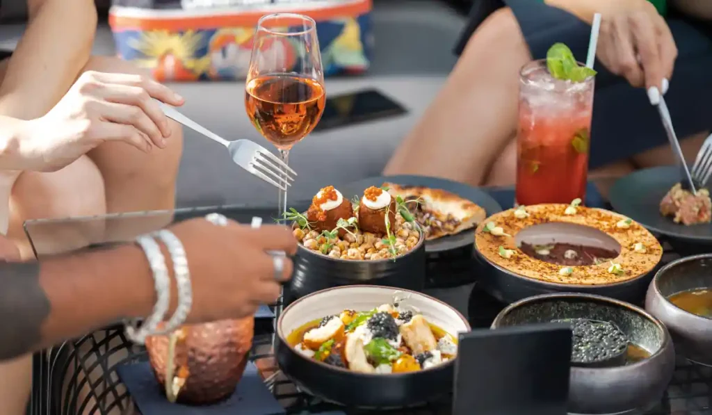 A table setting featuring a variety of dishes, including a bowl of chickpeas, a plate of appetizers, and several glasses of drinks. Three people are enjoying the meal, reaching for food with forks. A glass of rosé wine is prominently displayed.
