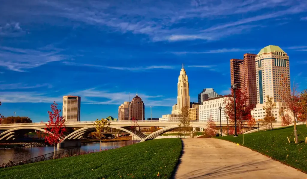 A vibrant cityscape of downtown Columbus, Ohio, featuring modern skyscrapers against a clear blue sky. A paved pathway runs alongside lush green grass and colorful autumn trees near a river, with an arched bridge in the background.
