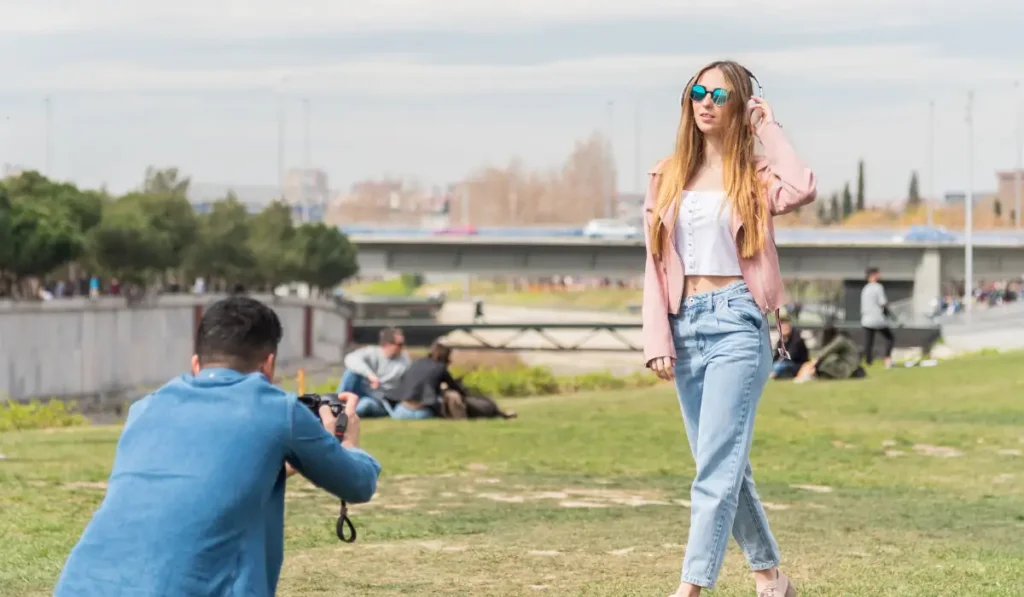 A woman poses outdoors in casual attire, wearing sunglasses and a pink jacket, as a photographer in a blue jacket takes her picture. They are in a park with grass and trees, and a bridge in the background.
