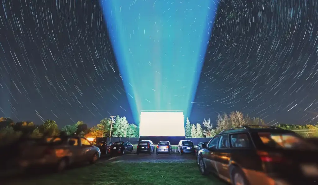 A timelapse photo of a drive-in movie theater at night. Cars are parked in rows facing a large screen projecting blue light upward. The night sky is filled with swirling star trails, creating a dynamic pattern over the scene.
