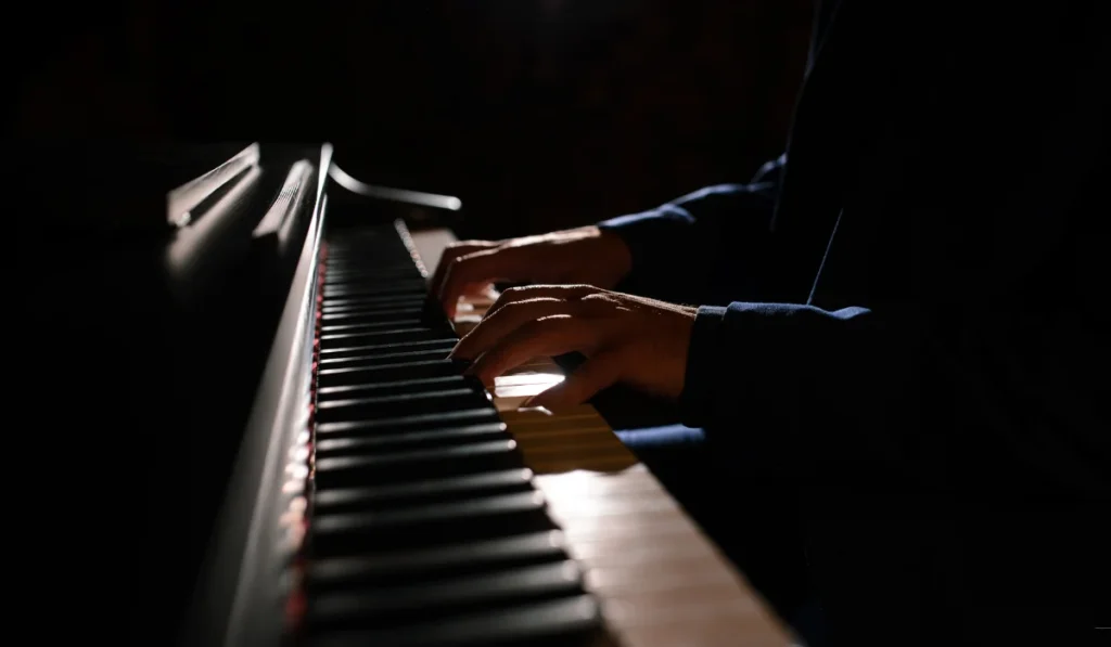 Hands playing a piano in a dimly lit setting, highlighting the keys and the musician's fingers. The scene conveys a sense of focus and musical expression.
