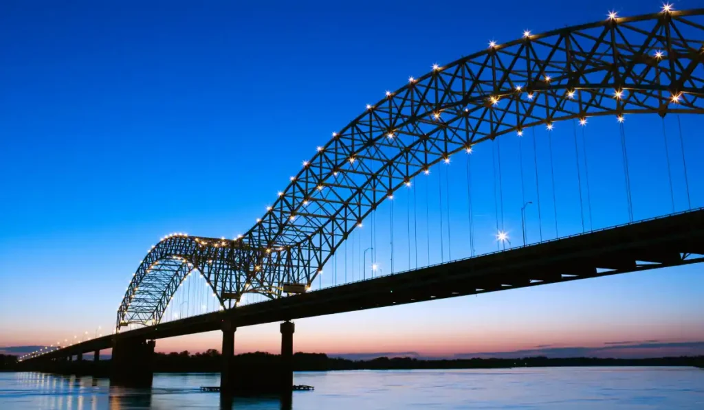 A large, illuminated arch bridge spans across a wide river at twilight. The sky transitions from deep blue to pink near the horizon. The bridges lights reflect softly on the water below.
