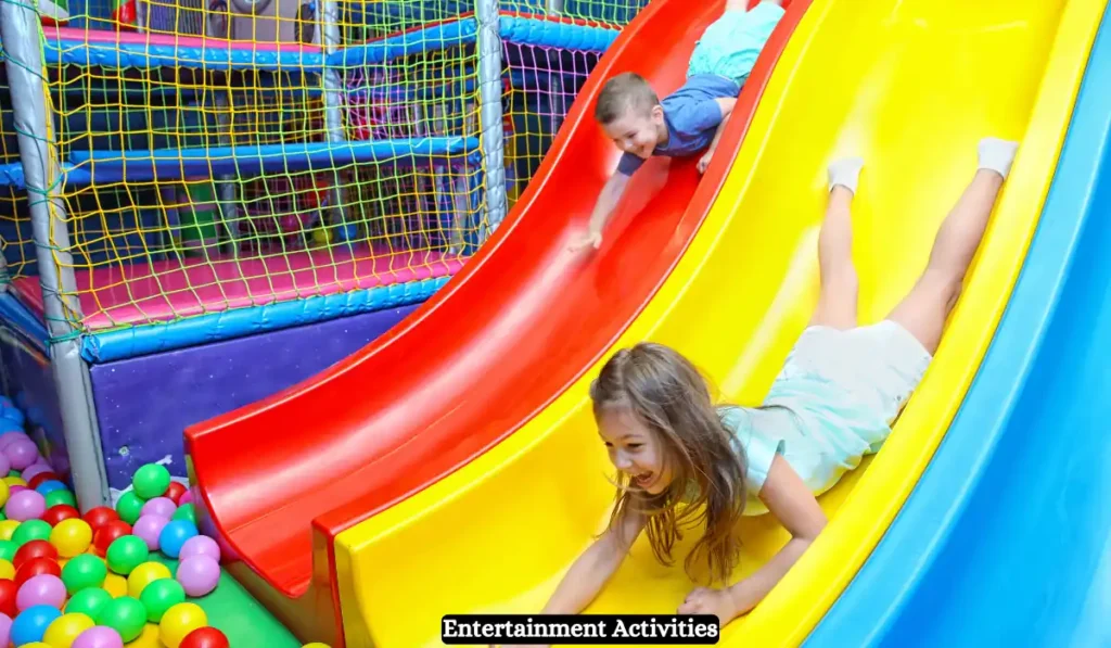 Children are happily sliding down colorful slides in an indoor playground with netted barriers and a ball pit at the bottom. The image captures the joy and excitement of the playful scene.
