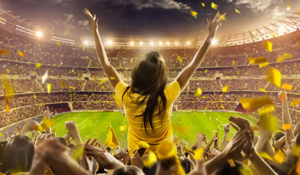 A woman in a yellow shirt stands with her arms raised, cheering in a packed stadium. The crowd around her is lively, with confetti in the air, and a brightly lit soccer field in the background.
