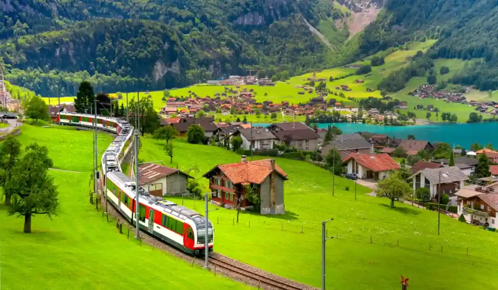 A passenger train travels through a lush green valley with scattered houses. In the background, a turquoise lake rests at the base of forested mountains. The sky is partly cloudy, adding depth to the picturesque landscape.
