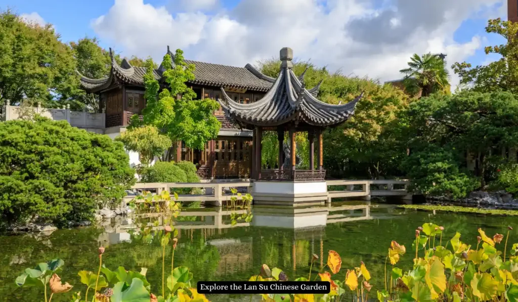 A serene Chinese garden with traditional architecture, featuring a pavilion with curved roofs and a lush green landscape. A pond with lotus plants reflects the structures and sky.
