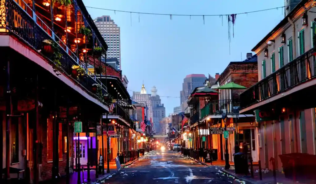 A vibrant street scene in New Orleans featuring illuminated buildings with balconies and streetlights under a dusky sky. The street is wet, reflecting the colorful lights, and skyscrapers are visible in the background.
