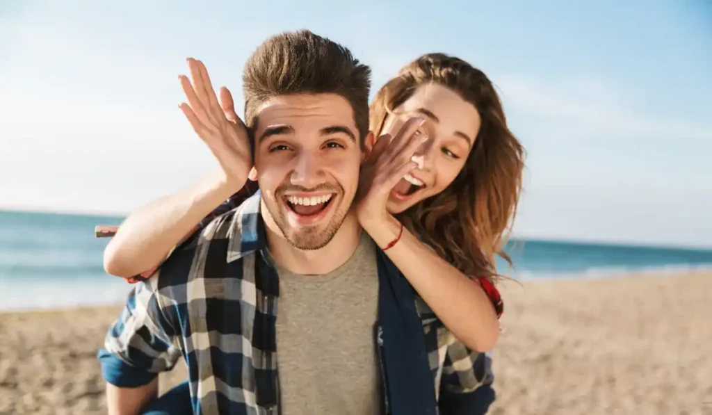 A smiling man carries a laughing woman on his back at the beach. They both seem joyful under the clear blue sky, with the ocean and sand in the background.
