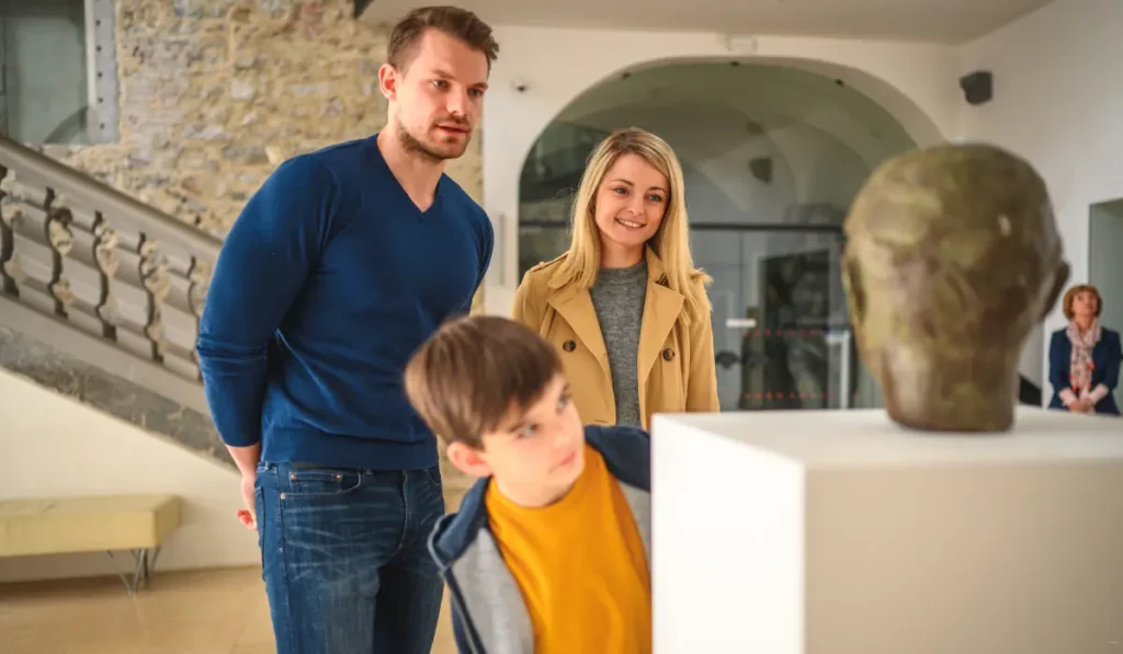 A family of three, consisting of a man, a woman, and a child, is observing a sculpture on display in a museum. The child leans in closely to look at the artwork. The room has bright lighting and a modern design.
