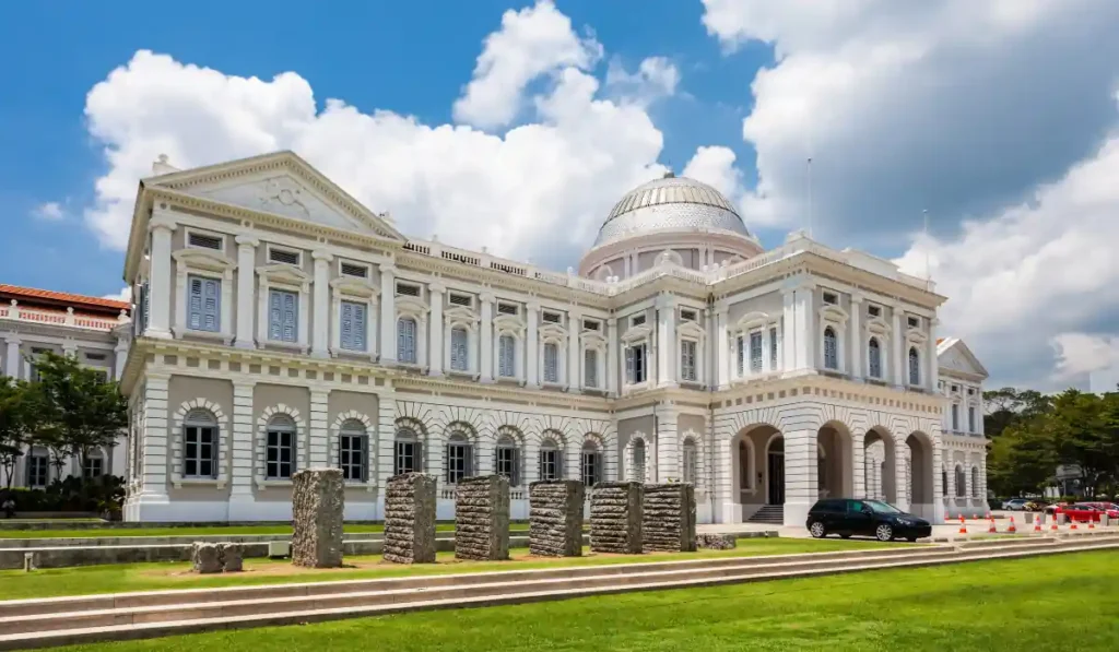 An elegant white colonial-style building with classical architectural features, including arched windows and a dome. It is surrounded by a well-maintained lawn and some stone blocks. The sky is partly cloudy.
