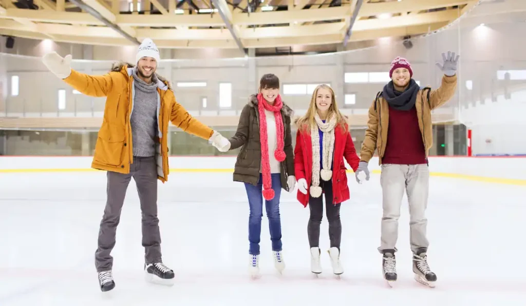 Four people holding hands and smiling while ice skating indoors. They are dressed warmly in winter clothes, including jackets, hats, and scarves. The ice rink has wooden beams and large windows in the background.
