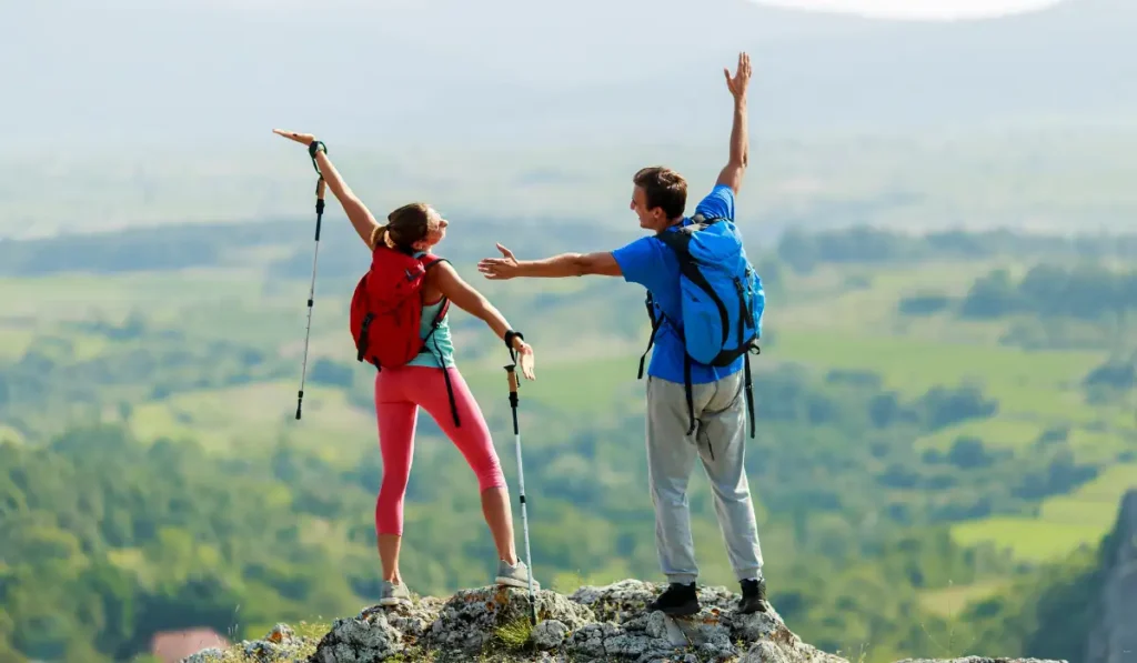 Two hikers with backpacks stand on a rocky cliff, arms raised in celebration. They overlook a vast, green landscape with rolling hills, enjoying a scenic view. Both hold hiking poles and appear joyful, embracing the adventure.
