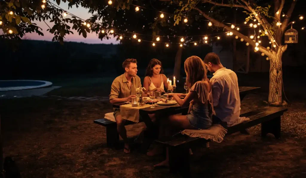 Four people sit at a wooden picnic table enjoying a candlelit dinner outdoors under string lights. The scene is set during twilight, creating a warm, inviting atmosphere. Trees and soft lighting add to the cozy ambiance.
