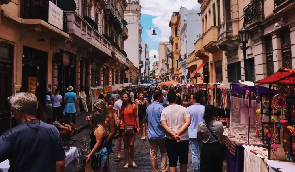 A bustling street market with people walking among colorful stalls displaying various goods. Historic buildings line the narrow street under a bright blue sky. Shoppers and vendors mingle, creating a lively atmosphere.
