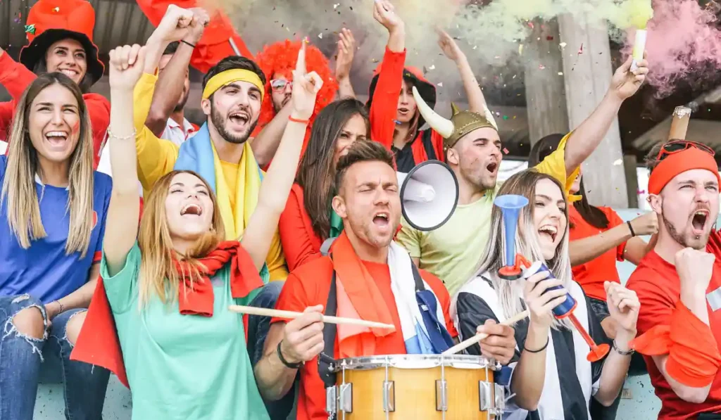 Enthusiastic sports fans cheer in a stadium, wearing colorful clothes and holding a megaphone and drum. Some have painted faces, and colorful smoke fills the air as they express excitement and support for their team.
