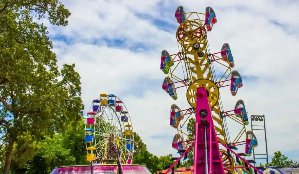 A vibrant amusement park scene featuring a colorful Ferris wheel and a towering thrill ride, surrounded by lush green trees under a partly cloudy blue sky.
