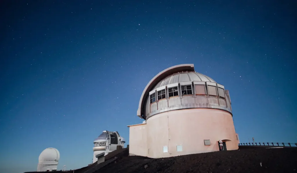 A clear night sky over a large astronomical observatory with multiple telescopes. The main building features a dome-shaped structure, and stars are visible in the deep blue backdrop.
