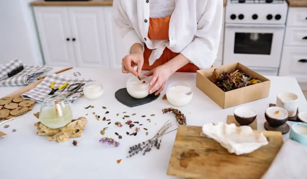 A person stands in a kitchen pouring wax into glass containers to make candles. The countertop is covered with dried flowers, herbs, tools, and a box of additional materials. The background shows white cabinets and a stove.
