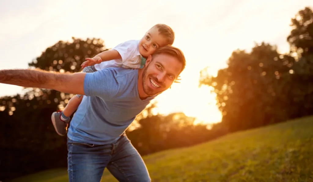 A smiling man in a blue t-shirt gives a little boy a piggyback ride in a sunlit park. The boy stretches his arms out, both appearing joyful against the backdrop of trees and a setting sun.
