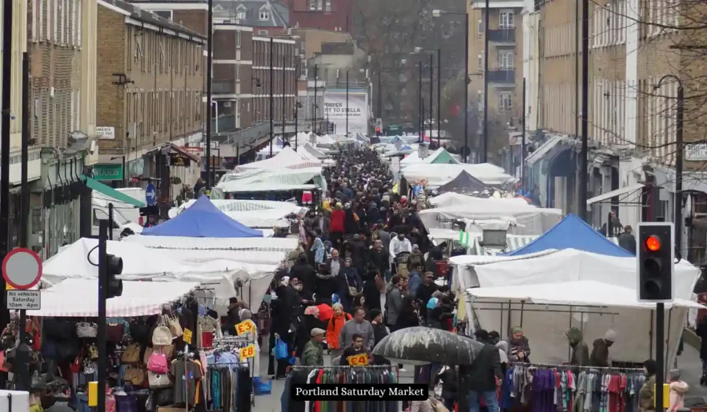 A bustling street market with numerous stalls covered by white and blue canopies. Crowds of people walk between the vendors, browsing goods. The market is set between tall brick buildings, and a "Portland Saturday Market" sign is visible near the front.
