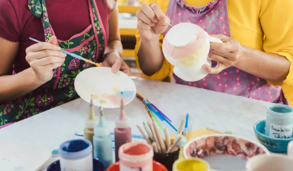 Two people painting ceramic items at a table. One holds a round plate, and the other holds a cup. Various paints and brushes are on the table. They are wearing aprons, and the atmosphere is colorful and creative.
