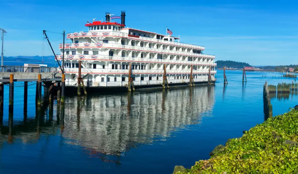 A large, multi-deck paddle steamer with an American flag is moored at a dock on a calm river. The blue sky and distant hills are reflected in the water. Green foliage lines the foreground.
