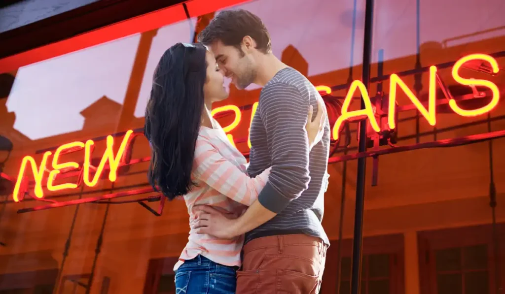 A couple embraces lovingly in front of a red neon sign that reads NEW ORLEANS. They smile at each other, enjoying a moment together. The signs glow reflects on the glass behind them. The setting suggests a lively, urban atmosphere.
