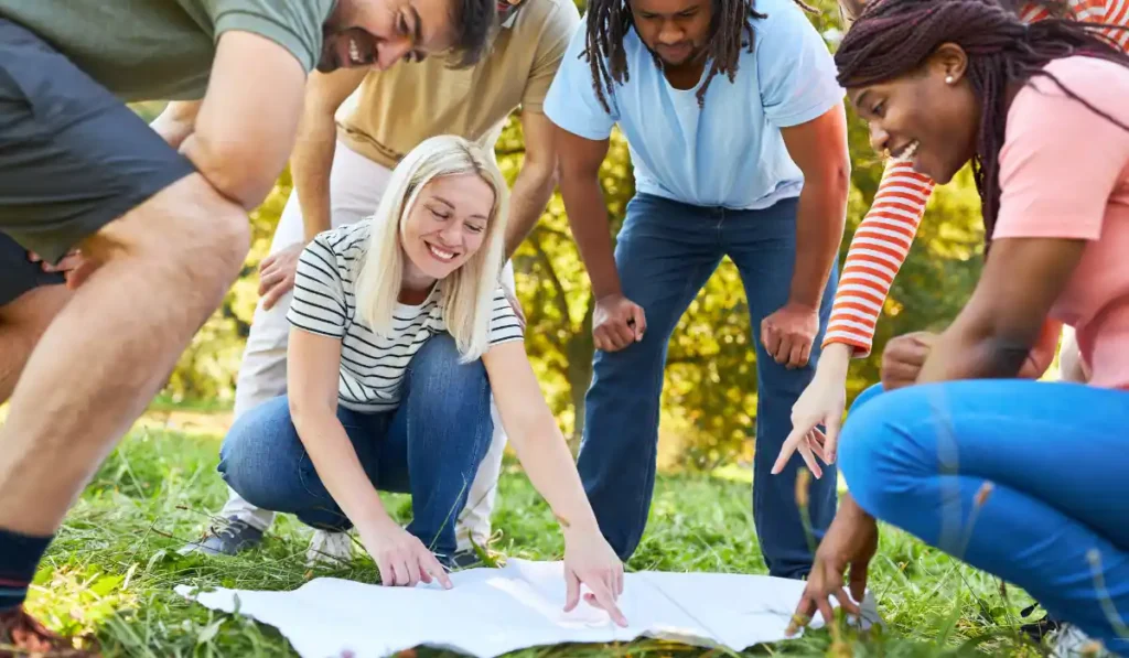 A group of people outdoors, bent over a large white sheet on the grass. They are smiling and pointing at the sheet, appearing engaged and happy. Sunlight filters through the trees, creating a lively and bright atmosphere.
