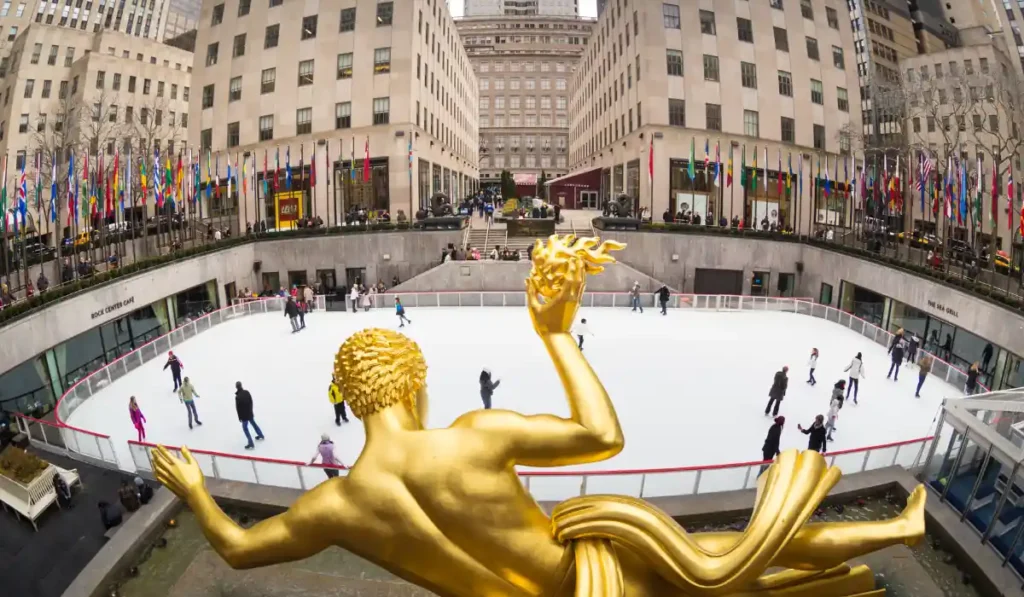 A view of the ice skating rink at Rockefeller Center in New York City, with people skating. The statue of Prometheus in the foreground is visible, and flags line the square. The surrounding buildings complete the urban scene.
