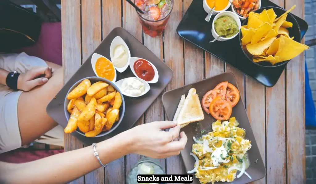 Top-down view of a wooden table with various snacks and meals, including seasoned potato wedges with different dips, a salad with tomatoes and toast, and a platter with guacamole and salsa alongside tortilla chips. Hands are reaching for food. Text reads "Snacks and Meals.