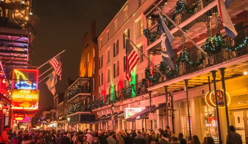 A lively, crowded street at night with buildings adorned in colorful lights and decorations. Flags hang from balconies, and the bright glow of neon signs illuminates the scene, creating a festive atmosphere.
